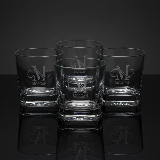 Four custom-engraved square heavy-base whiskey rocks glasses featuring a personalized monogram and name design (M with Morgan underneath), displayed together on a reflective surface