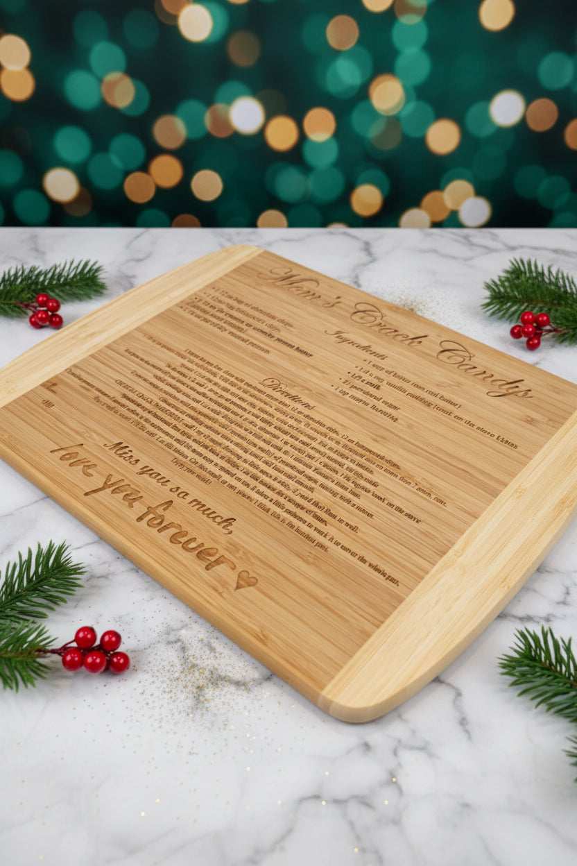A rectangular bamboo cutting board featuring a custom-engraved Christmas heirloom recipe and a handwritten 'love you forever' message, styled on a white marble surface with pine branches, red berries, and a festive green-and-gold bokeh background.