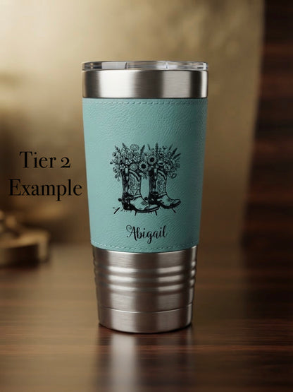 A stainless steel insulated tumbler with a light blue leatherette sleeve, featuring a black graphic of cowboy boots filled with wildflowers and the name 'Abigail' engraved below. The tumbler is shown on a wooden table with a warm, blurred background, and the text 'Tier 2 Example' is visible on the left. 