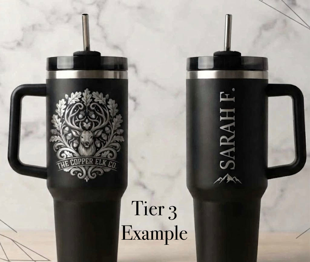 A black insulated tumbler with handles duplicated to show the front and back side by side on a light marble background. The left tumbler (front side) has a silver elk  head and ornate tree design with the text 'THE COPPER ELK CO. The right tumbler (back side) has the name SARAH F engraved vertically with a mountain graphic underneath. Tier 3 Example is a text overlay on the image.