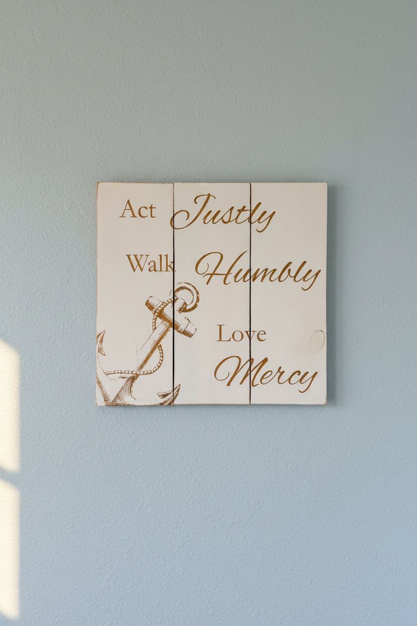 A rustic white wooden sign custom laser engraved with text reading 'Act Justly Walk Humbly Love Mercy' and an anchor illustration, mounted on a light blue wall. 