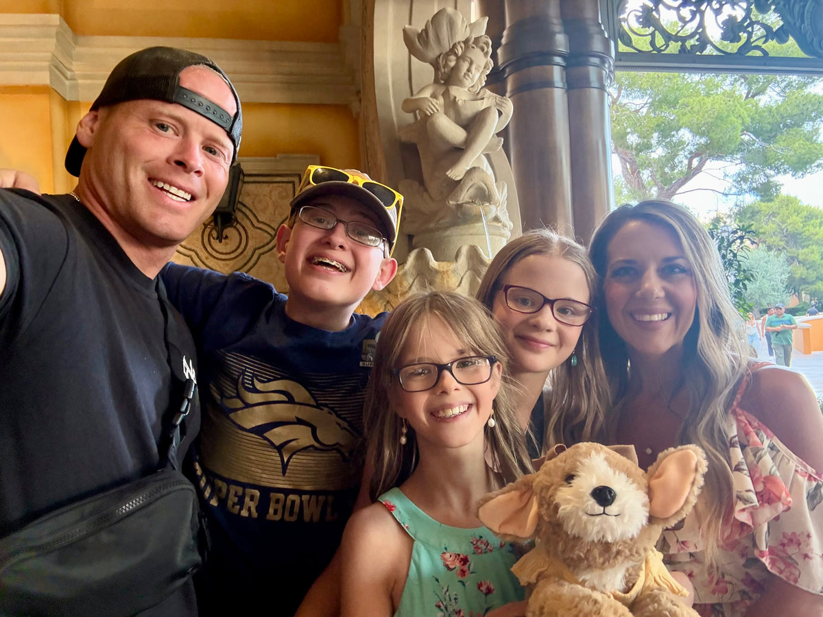 A family of five posing together in front of an ornate architectural background with a statue (outside Bellagio hotel in Las Vegas). The group includes a man taking the selfie, a boy in a Denver Broncos shirt, a girl holding a plush dog toy, a girl with magenta glasses, and a woman in a white and pink floral dress.