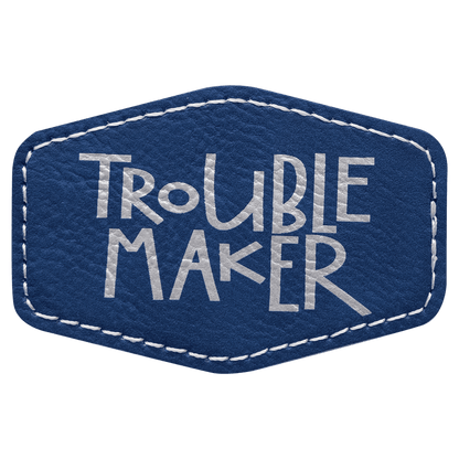 A blue leatherette hexagon patch with stitched white edges, featuring the text "TROUBLE MAKER" in a distressed silver-gray block font.
