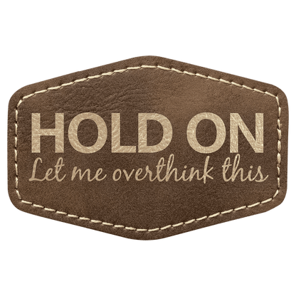 A brown leatherette hexagon patch with stitched edges, featuring the text "HOLD ON Let me overthink this" in a mix of block and script lettering.