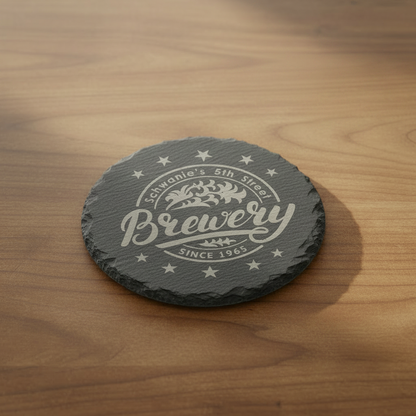 A stack of six circular natural slate coasters, featuring rough, hand-chiseled edges and a laser-engraved brewery logo in the center.