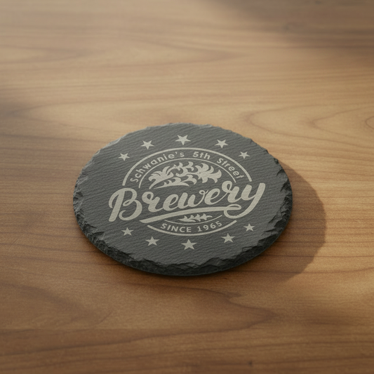 A stack of six circular natural slate coasters, featuring rough, hand-chiseled edges and a laser-engraved brewery logo in the center.