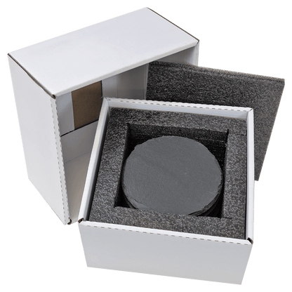 A high-angle studio shot of a white cardboard box partially open to reveal six round, black slate coasters neatly stacked inside protective black foam packaging.
