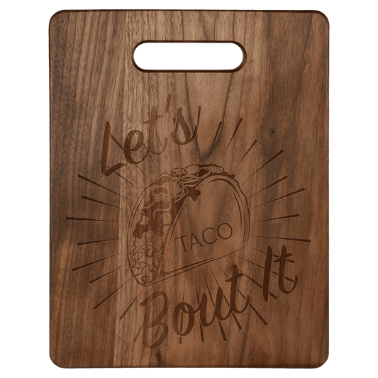 Rounded rectangular walnut cutting board with integrated handle, featuring a custom laser-engraved taco graphic and the text 'Let's Taco Bout It'